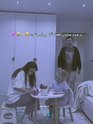 A family scene is captured in a room with warm lighting. The image features a woman and a young girl sitting at a small white table. The little girl is drawing with crayons while the woman is looking at a small cup with pens. A man stands in the background, looking like he is observing the family from behind.