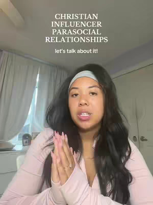 A woman with long, dark hair wearing a light pink top, and a headband. Her hands are clasped together in a prayer-like gesture. Behind her are curtains and a wall.