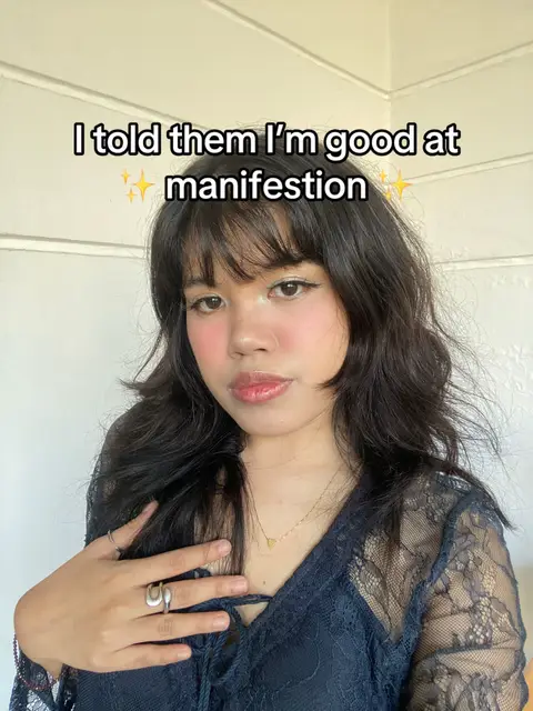 A selfie of a young woman with long, dark hair and bangs, looking directly at the camera. She's wearing a dark lace top and a necklace with a small heart pendant. Her makeup includes blush and lip gloss. Text overlay reads: 'I told them I'm good at manifestation'.