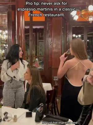 The cover image shows three women in what appears to be a classic French restaurant. One woman is standing and talking, wearing a white top and checkered pants. Another woman is seated at the table. A third woman is standing and drinking from a glass, wearing a black dress with an open back. The restaurant has dark wood paneling and ornate lighting.