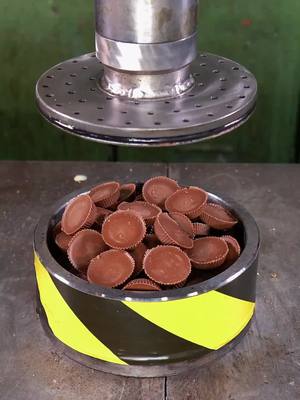 A close-up shot of a metal cylinder filled with numerous peanut butter cups. Above, a metallic press is positioned to descend onto the cups. The cylinder has a black band with yellow diagonal stripes, and the background is the gray surface that the cylinder is resting upon.
