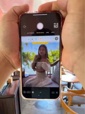 A person is holding a phone that is displaying a photo of a woman sitting in a cafe. The hands holding the phone are visible in the foreground. The background of the photo on the phone shows the cafe setting with a woman and tables.