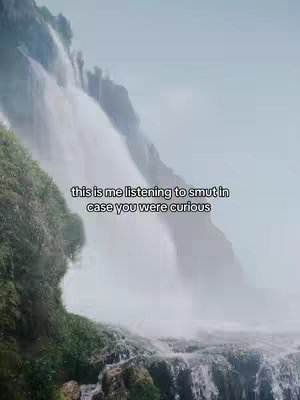 A scenic shot of a cascading waterfall. The water plunges down a rocky cliff face, creating a misty veil. Lush green vegetation clings to the cliff edges. A text overlay at the bottom reads, 'this is me listening to smut in case you were curious'.
