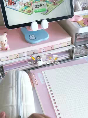 The image shows a well-lit desk setup with a tablet and a notebook, hinting at a study or work environment. The color scheme is soft, with pastel pinks and whites dominating the scene. In the foreground, a notebook with grid paper and a pencil case are visible. In the background, a tablet is propped up, displaying an interface. There are also various desk accessories, including a white rabbit figurine and a bunny-shaped tablet stand.