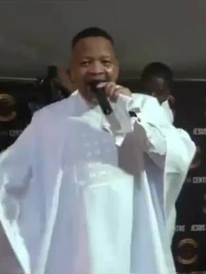A man in a white robe speaks into a microphone, with a blurry background hinting at an event or service. The color scheme is bright, and the composition is focused on the man.