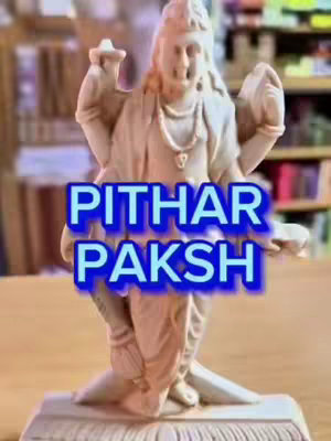 A statue of a Hindu deity is displayed, placed on a wooden surface. Text overlayed on the image in a blue outline reads 'PITHAR PAKSH'. The background is blurred, featuring indistinct objects.