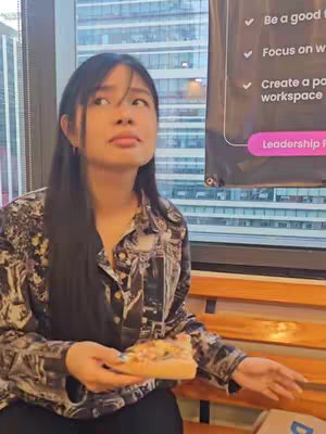 A young woman with long dark hair, wearing a patterned shirt, looks up and to her right with a slight expression of surprise. She is holding a slice of pizza, with a building and a chart/sign visible in the background.