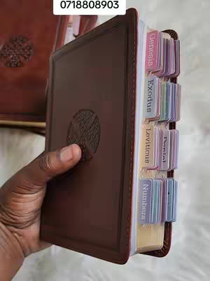 A close-up shot displays a brown leather-bound Bible with colored tabs, held by a hand. The tabs show the text indicating the book of the bible.  Another leather-bound object is visible in the background against a white surface.