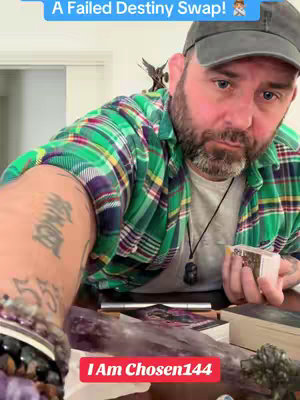 A medium shot shows a man in a green plaid shirt, a baseball cap, and a beard. He appears to be holding tarot cards, with crystals and other items on the table in front of him.