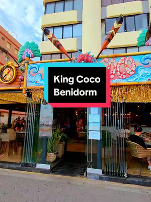 The cover image showcases the exterior of a restaurant called King Coco in Benidorm. The restaurant has a colorful facade with decorative elements and a sign that reads 'King Coco Benidorm'. A black and teal rectangle overlaid with white text indicating the name and the location of the business. Through the glass doors and windows, patrons can be seen. The overall composition suggests a lively and inviting atmosphere.