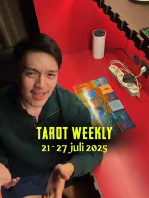 A man sits in front of a bright red surface, possibly a table or counter, with a semi-circular black border. A white cylinder object, possibly a speaker, and a strip of a magazine are on the surface. The man is smiling. Text on the image identifies the video's content as a 'Tarot Weekly' for the week of July 21-27, 2025.