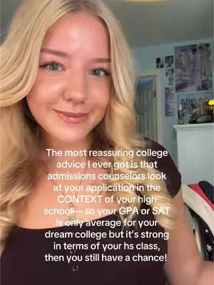 A selfie-style video features a young woman with blonde hair, blue eyes, and a subtle nose ring. She is positioned in front of a lightly colored wall, potentially in a well-lit bedroom, and is centered in the frame from the chest up. The background elements include a door, posters, and a white dresser, indicating a personal space. The woman is looking at the camera with a warm and inviting expression, dressed in dark-colored top. A text overlay in a sans-serif font is visible on the image. The overall composition is bright and emphasizes the subject against a backdrop of neutral tones.