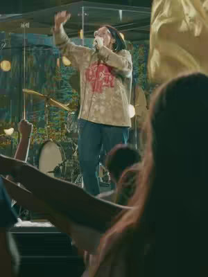 A live music performance scene at a concert, with a man on a stage singing into a microphone and extending his arm towards the sky. A multitude of audience members are in focus in foreground, arms raised in worship or praise. A drum set is partially visible behind the singer. The background has some creative color, with some golden elements in it.