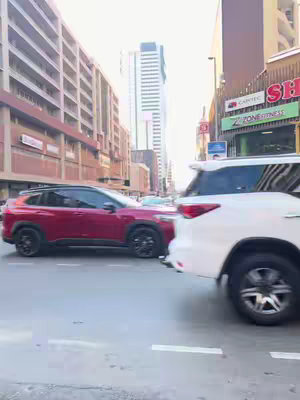 A street scene with a red car in motion on the left and a white SUV on the right side, set against a backdrop of buildings and a clear sky. The composition captures a moment in an urban environment.