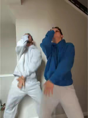Two young men in casual attire are dancing with their arms in the air, set against a neutral-toned wall, creating a playful and energetic visual.