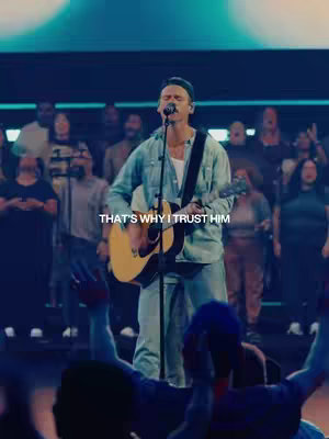 A live performance scene with a man singing and playing a guitar on stage. He is wearing casual attire and is in front of a crowd. The background is blurred, with spotlights.