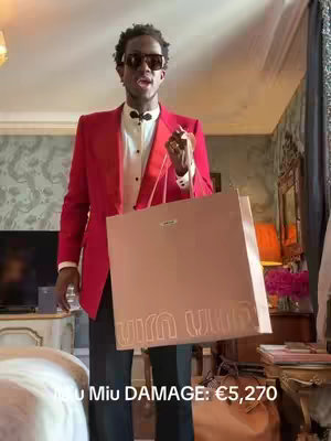 A man in a red blazer, white shirt, and black pants stands in a room, holding a large shopping bag. He's wearing sunglasses and a bow tie. The shopping bag is peach-colored with a logo on it, and he's also holding a small bottle. The text 'Miu Miu DAMAGE: €5,270' is at the bottom.