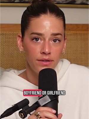 A woman with light skin and light brown hair in a bun is seated at a desk or table facing the camera. She is wearing a white hoodie and is holding a microphone. The background is slightly blurred, but appears to be a room with some sort of woven material on the wall.