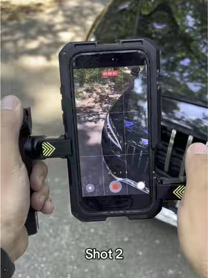 An image of a person holding an iPhone in a special mount in front of a car. The phone screen displays a recording interface, capturing what appears to be the front of the car. The car seems to be a Mercedes-Benz. The hands are holding the mount with the phone attached. The layout seems designed to showcase the phone's video capabilities while filming a car.