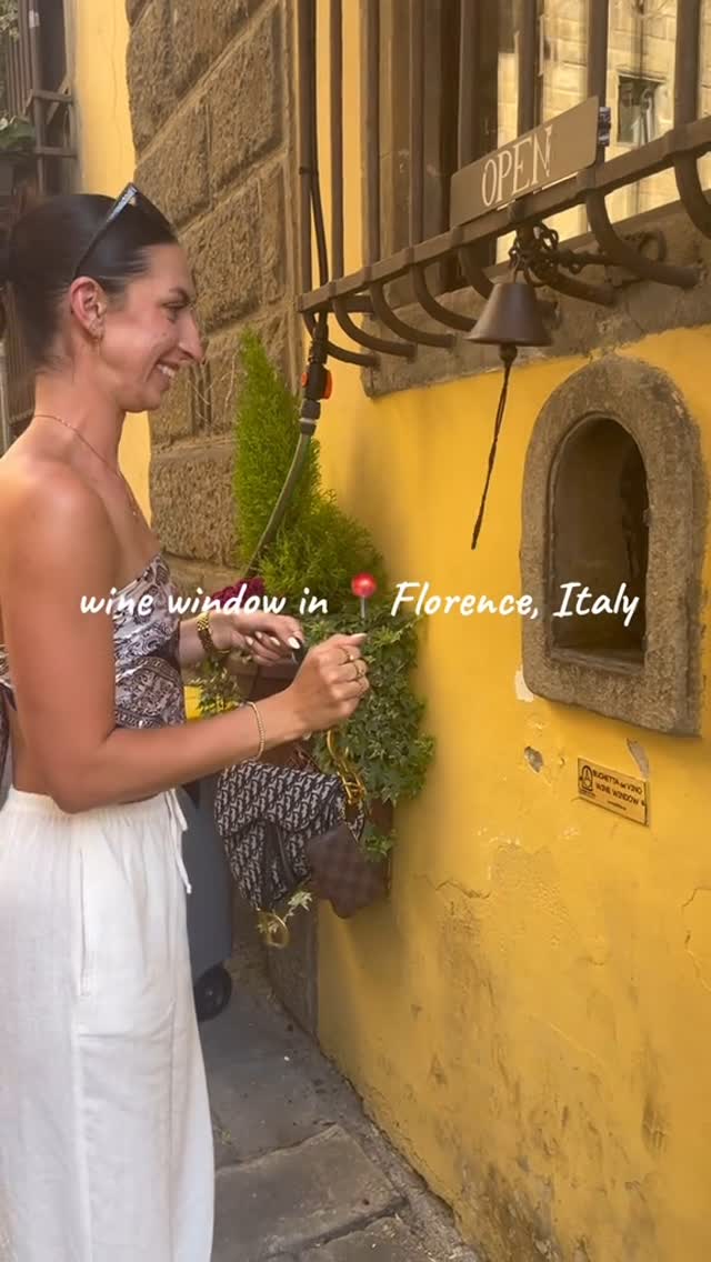A woman stands beside a yellow building with a small, arched window, likely a wine window. A small bell hangs next to the window. Text overlay says "wine window in Florence, Italy". The woman is wearing a patterned top and white pants, and appears to be interacting with the bell or window. The setting is outdoors with stone brick visible on the building. The background has a plant in a decorative container.