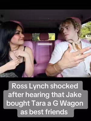 A person in a white shirt, sits behind the wheel of a car, talking to another person in the passenger seat. The interior of the car has pink seats.