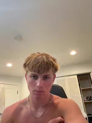 A young man with light skin and short, tousled blond hair is centered in the frame. He is shirtless, wearing a silver chain necklace. The background includes a white ceiling with recessed lights, a white storage cabinet, and the corner of a room with doors and a white trim. The lighting is soft.