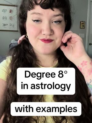A selfie-style shot featuring a woman with long, wavy dark hair, parted on the side. She gazes towards the camera with a soft smile, her hand gently touching her face near her cheek. Dressed in a yellow top, she has red lipstick, dark eyeliner, and a small floral tattoo on her arm. Overlaid on the image is a white rectangular text box with black text saying 'Degree 8° in astrology with examples'. The background includes parts of a room with a framed image.