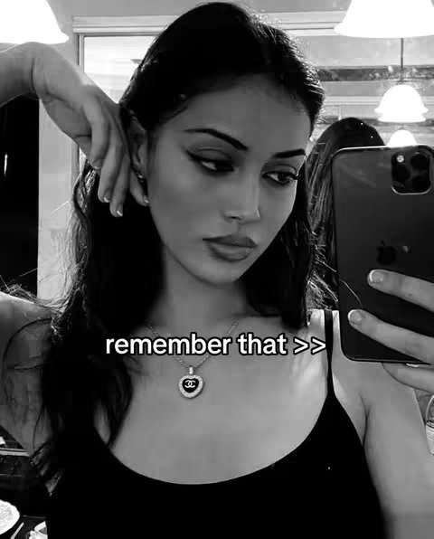 A grayscale selfie-style image featuring a young woman gazing downwards while holding a smartphone, possibly taking a photo. She wears a black tank top, and a heart-shaped pendant necklace. The overall tone is chic, with focus on the woman's face and expression. A hand is held up in the background.