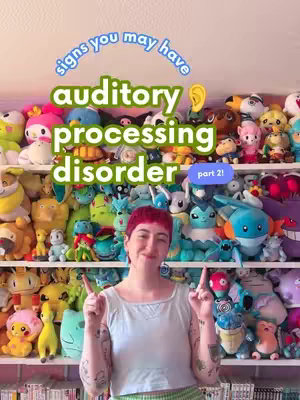 The cover image shows a person with red hair, framed from the chest up, pointing at the viewer with both index fingers. They are in front of a backdrop of shelves packed with colorful stuffed animals. Text overlays include "signs you may have" in a curved blue font on top, the phrase "auditory processing disorder" in the center, and "part 2!" in a small purple box. The image uses a variety of bright colors, primarily blues, greens, and pinks, creating a playful and informative feel.