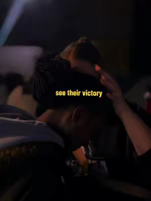 The image shows a dimly lit scene with two figures in a tight embrace or hug. The focus is soft, with the individuals' faces partially obscured by shadow and the presence of another person's hand partially visible. A text overlay in yellow, reads 'see their victory'. The overall composition leans heavily on the emotional aspect, with the contrast of light and shadow.
