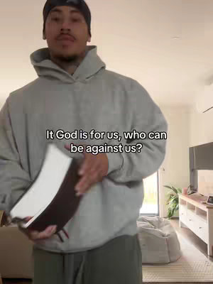 A man in a grey hoodie and a black beanie holds a book in a home setting.