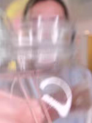 The image is blurry. The main element is a hand close to the camera's lens, partially obscuring the face of a person in the background. The hand appears to be holding something, possibly a glass, reflecting light and creating a hazy effect. The composition is somewhat blurred, giving a sense of movement or immediacy . The overall color palette seems to be soft and muted.