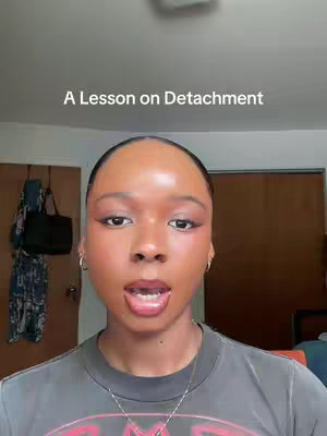 A selfie-style shot featuring a dark-skinned person with short hair, centered in the frame. The person has a neutral expression, looking directly at the camera. Behind them, a plain wall backdrop is visible, with a door and some hanging items on the left side. The lighting appears to be from an indoor source.