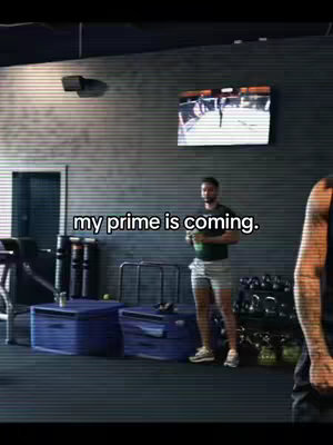 A man in workout clothes holding a weight stands in a gym. The background has a TV screen and workout equipment.