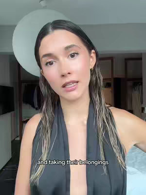 A woman with long, wet, dark hair and wearing a black, sleeveless outfit appears to be addressing the camera. The background is a blurred room with a large, circular overhead light. Text is overlaid at the bottom of the frame.