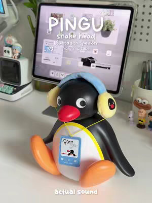 A photograph of a desk setup featuring a Pingu-shaped Bluetooth speaker wearing blue headphones, situated in front of a tablet displaying a music app interface. The tablet and the speaker are placed on a white desk with other decorative objects and writing utensils. A window with a green plant is visible in the background. The overall composition is clean, bright, and kawaii-inspired.