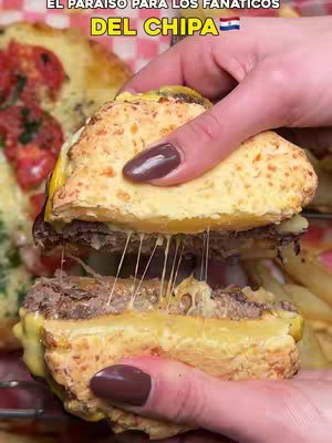 A close-up shot of a burger made with a chipa bun, being held by hands with polished nails. The burger is cut in half, with cheese stretching between the layers. The background shows other food items and is slightly blurred.