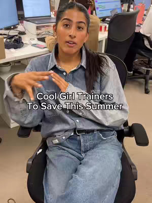 A woman in a denim shirt and jeans is seated in an office chair, gesturing with her hands. Text overlayed reads: Cool Girl Trainers To Save This Summer.
