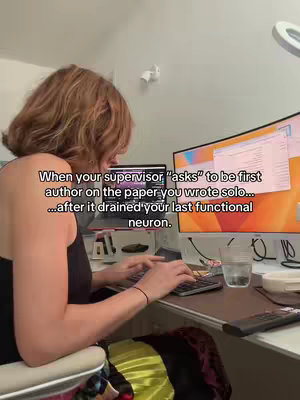 A woman is seated at a desk working on a computer and laptop. Overlay text is on the screen of a laptop.