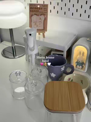 The image showcases a collection of home decor items arranged on a white surface. There are glass containers with lids, a blue floral mug, a wooden-topped container, and a bedside lamp with a bunny figurine. Overlay text 'Shein home haul' with heart emojis.