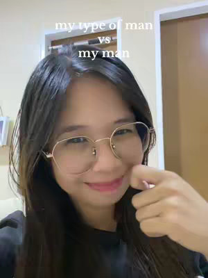 A selfie of a woman with mid-length dark hair, wearing gold-rimmed glasses, and a dark-colored top. She is smiling slightly, with her right hand near her mouth, and the background shows a room with a white door and a glimpse of a wall art or picture. Text overlay on the top shows 'my type of man', 'vs', and 'my man'.