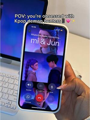 A person holding an iPhone displaying animated scenes from Kpop. The phone screen showcases an ongoing WhatsApp audio call with a call in progress, featuring images of animated characters. The background gives glimpses of a laptop with a white keyboard and other desktop elements.