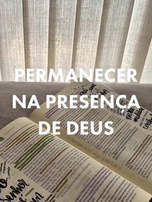 The image features an open Bible with highlighted passages, set against a backdrop of softly lit, vertical blinds. The text overlay in large white font reads, 'PERMANECER NA PRESENCA DE DEUS,' translating to 'Remain in the presence of God'. The composition is warm and focused, suggesting a devotional setting.