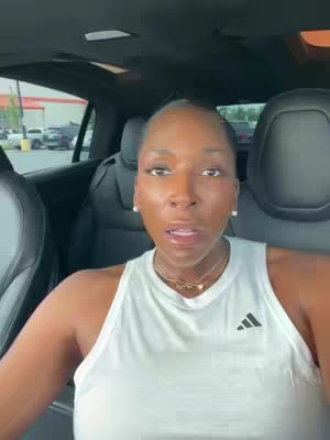 A woman is seated in the driver's seat of her car, looking directly at the camera. She is wearing a white athletic tank top, and a delicate necklace. The car interior is black, with the seats and dashboard visible. A blurry outdoor scene is visible behind the car.