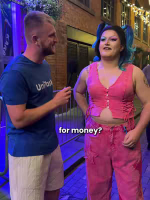 Two people standing in the street, a man in a blue t-shirt and white shorts on the left and another person in a pink outfit on the right. The person in pink is standing with their hands on their hips, and the man is holding a phone and pointing towards them. Text is overlaid on the image.