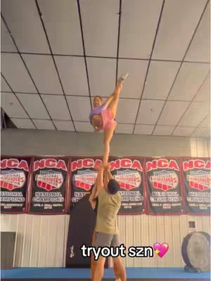 The image shows a cheerleading stunt in progress. Two people are supporting a flyer who is in a split in mid-air. The background includes banners, a plain wall and a ceiling with lights.
