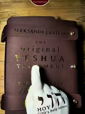 The image shows a close-up of a leather-bound book, likely a Bible, with gold lettering. A hand wearing a foam finger points towards the book. Behind the book is a wooden surface. The book is brown with a gold inscription reading "ALEKSANDR LASTIVKA" at the top and "The original YESHUA TESTAMENT" in the center. The foam finger has the words "הוי  HOY  GOD's holy letters" in Hebrew and English. In the upper part of the image is a small logo.