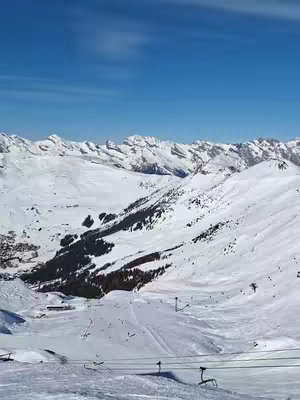 A scenic view of snowy mountain slopes under a blue sky. The foreground shows a ski slope with ski lifts, while in the distance, other snow-covered mountains rise.