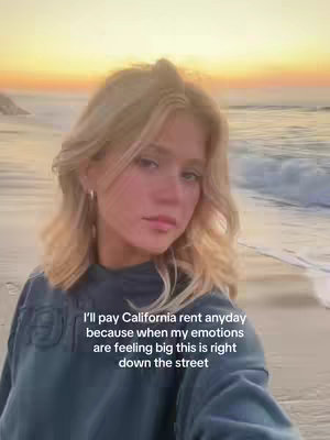 A young woman with long blonde hair poses on a beach at sunset. The sky is shades of orange and yellow, the ocean is a calm blue, and the sand is a pale tan. The woman is wearing a teal-blue long-sleeved shirt and gazing softly at the camera. Text is overlaid on the bottom of the image.