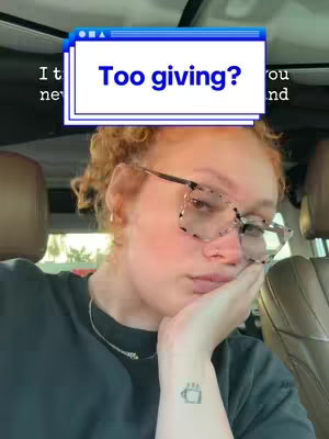 A selfie taken inside a car, featuring a woman with reddish-brown curly hair, wearing glasses and a black t-shirt. She is touching her cheek with her hand, glancing downwards with a thoughtful expression. A text overlay asks 'Too giving?'.
